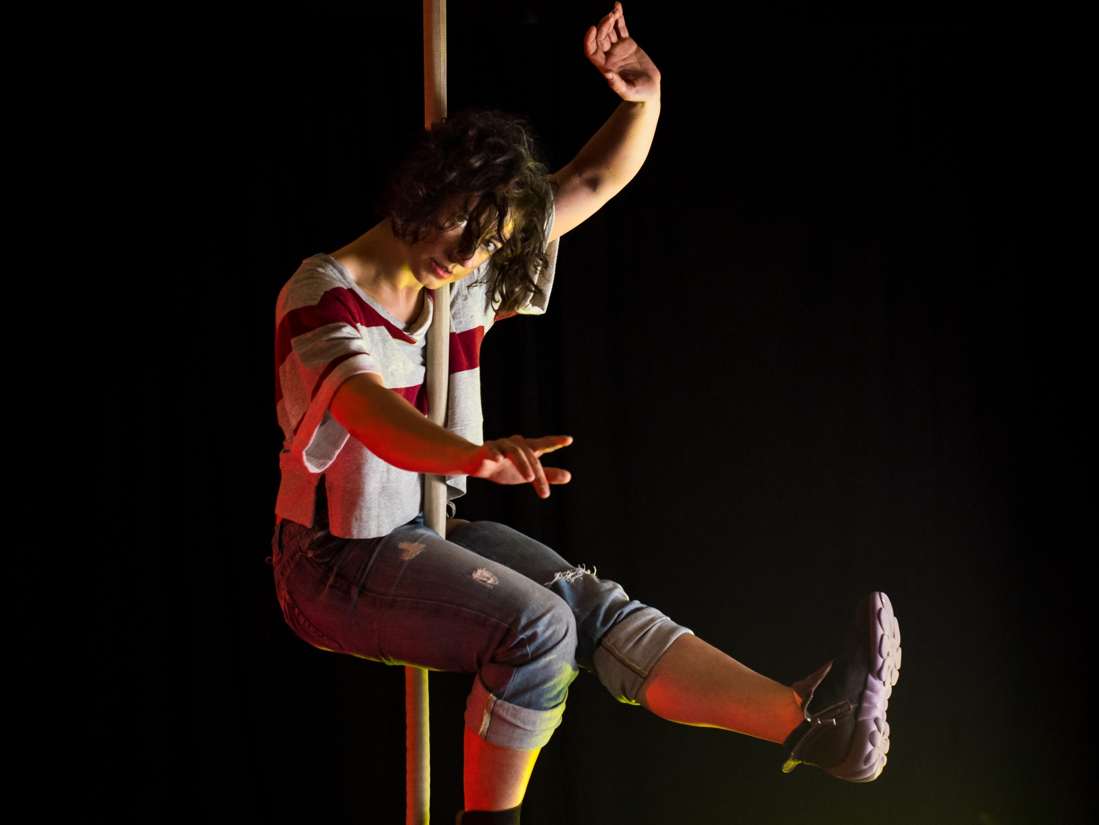 A woman suspended by a rope in a sitting position. She's wearing torn jeans and a bag red and gray shirt. Here eyes look into the camera and her arms reach haphazardly into space.