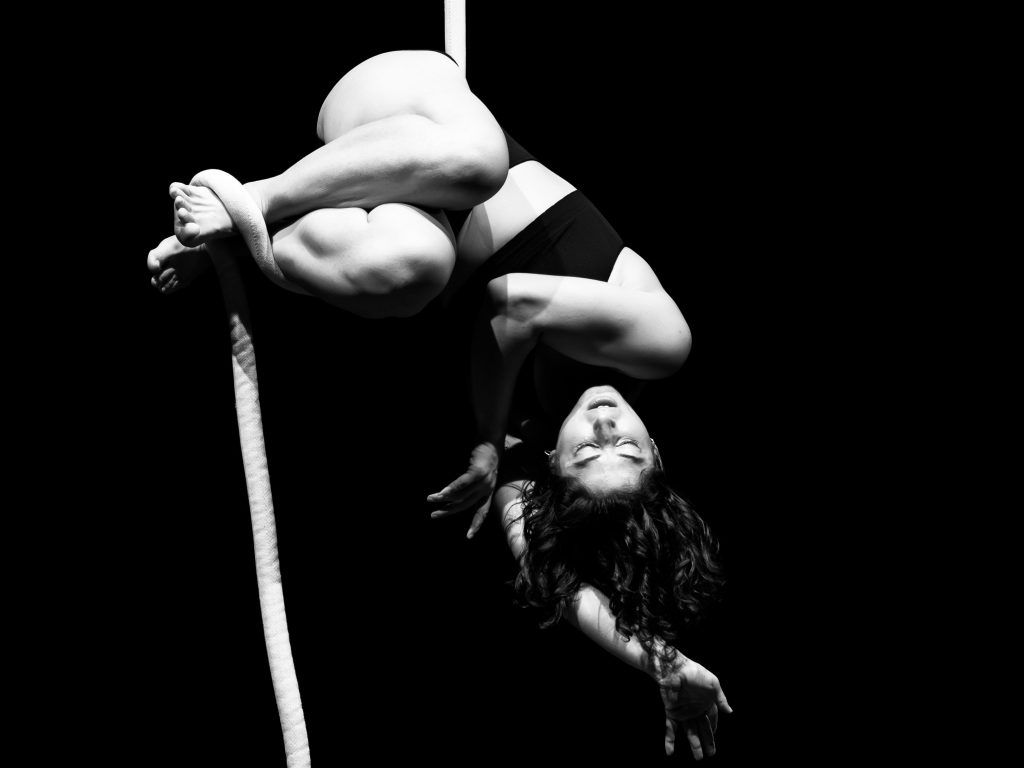 Black and white. A woman hangs sideways curled up in a ball with eyes closed on a white rope. 